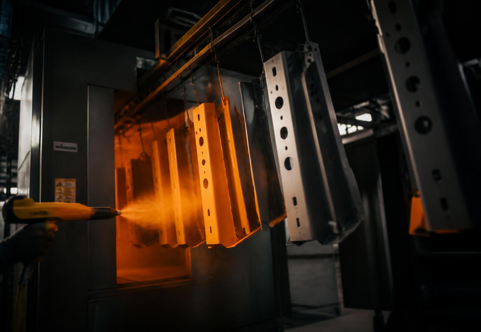 Powder coating spray gun applying orange powder to steel parts on a conveyor line
